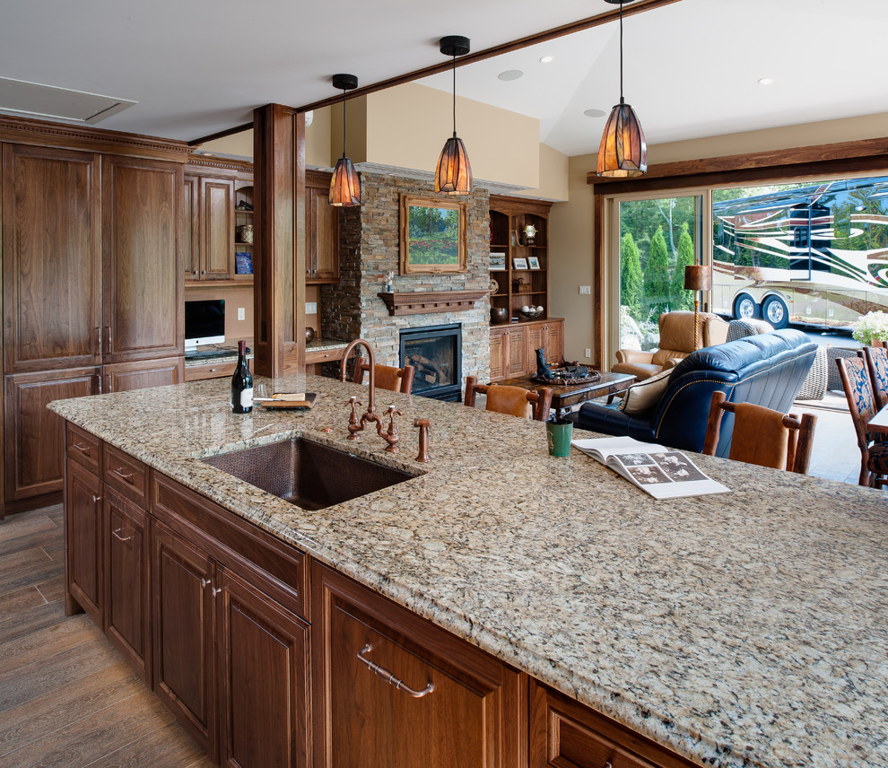 Example of a large mountain style u-shaped medium tone wood floor and brown floor open concept kitchen design with an undermount sink, raised-panel cabinets, medium tone wood cabinets, granite countertops, beige backsplash, travertine backsplash, paneled appliances and an island