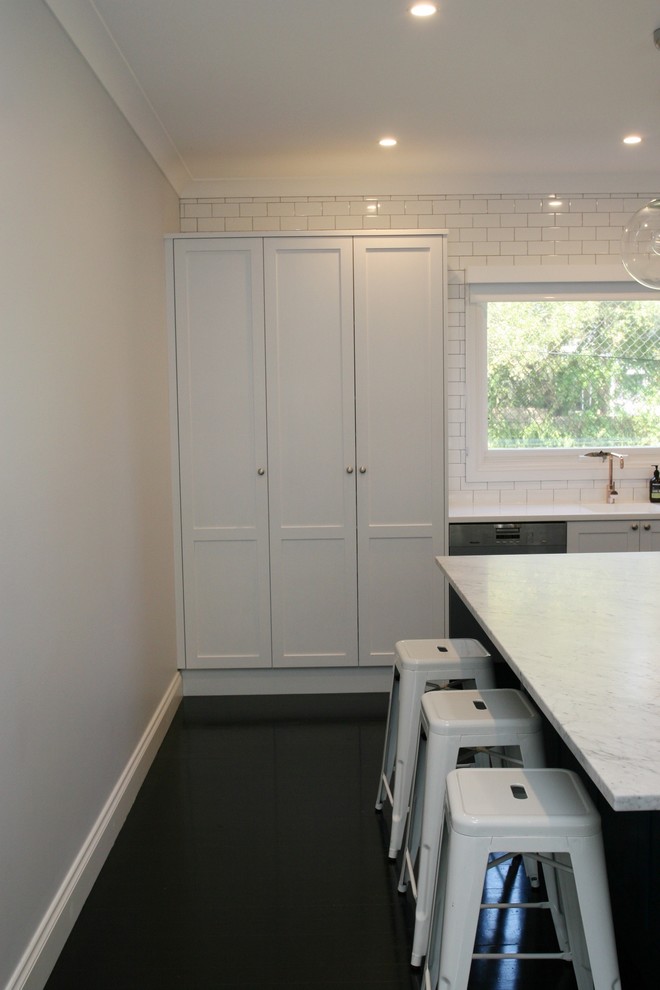 Large traditional l-shaped open plan kitchen in Sydney with an integrated sink, shaker cabinets, white cabinets, marble benchtops, white splashback, stainless steel appliances, with island, subway tile splashback, painted wood floors, black floor and white benchtop.
