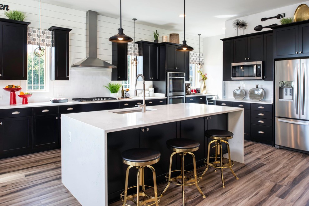 Photo of a country l-shaped kitchen in Cincinnati with an undermount sink, black cabinets, white splashback, subway tile splashback, stainless steel appliances, with island, brown floor and white benchtop.