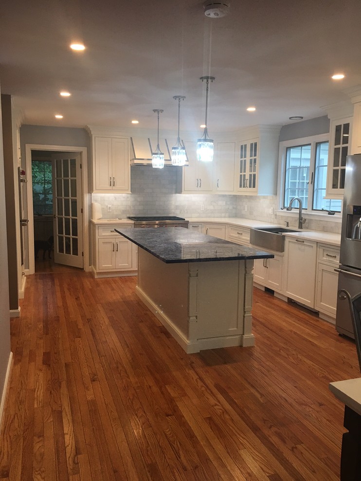 Mid-sized transitional l-shaped medium tone wood floor and brown floor kitchen photo in Boston with a farmhouse sink, shaker cabinets, white cabinets, granite countertops, gray backsplash, subway tile backsplash, stainless steel appliances and an island