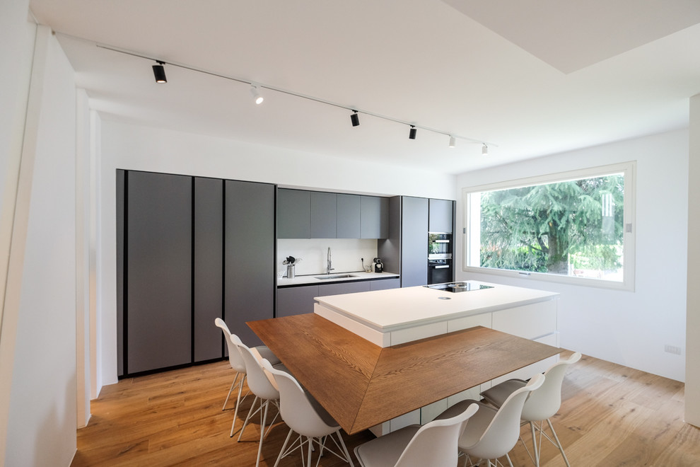 Inspiration for a contemporary kitchen in Milan with an undermount sink, flat-panel cabinets, grey cabinets, white splashback, black appliances, light hardwood floors, with island and white benchtop.