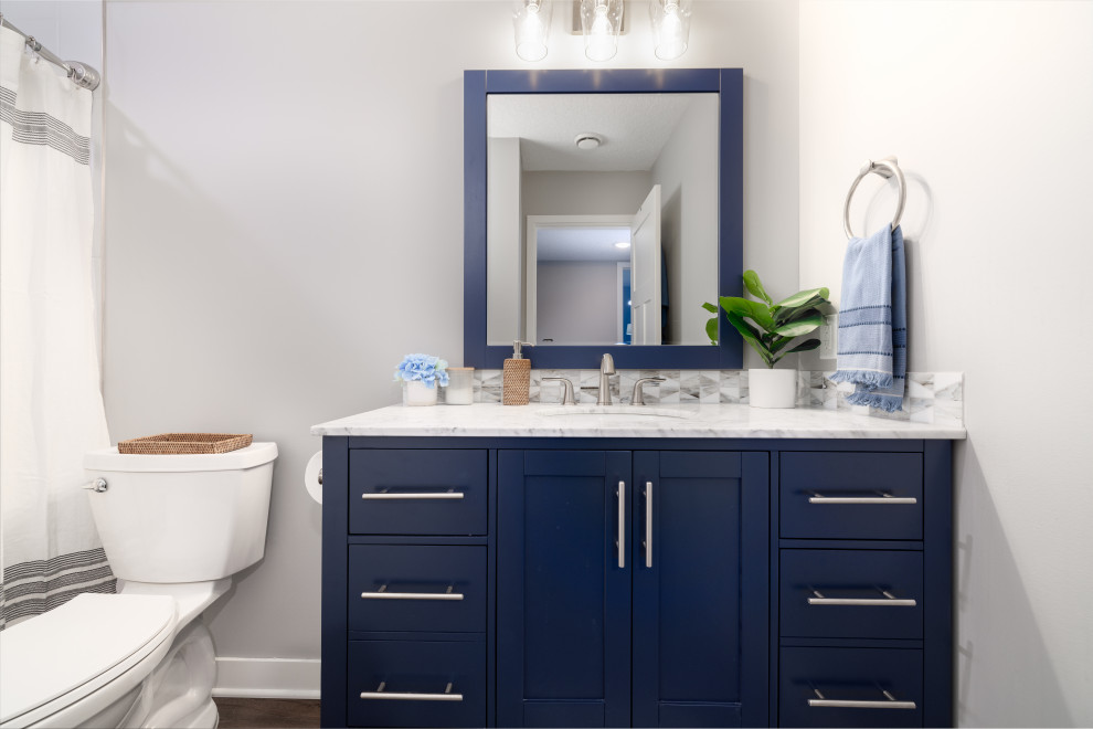 This is an example of a mid-sized transitional 3/4 bathroom in Other with shaker cabinets, blue cabinets, an alcove shower, a one-piece toilet, white tile, porcelain tile, grey walls, vinyl floors, an undermount sink, engineered quartz benchtops, brown floor, a shower curtain, white benchtops, a single vanity and a freestanding vanity.
