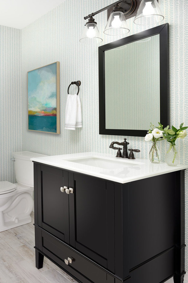 This is an example of a mid-sized beach style powder room in Toronto with recessed-panel cabinets, brown cabinets, a two-piece toilet, multi-coloured walls, laminate floors, a drop-in sink, engineered quartz benchtops, grey floor and white benchtops.