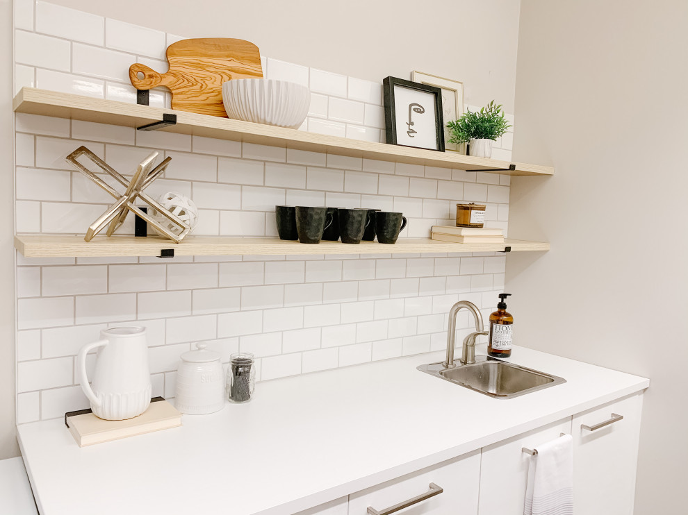 Small transitional single-wall wet bar in Vancouver with a drop-in sink, flat-panel cabinets, white cabinets, laminate benchtops, white splashback, subway tile splashback, vinyl floors, brown floor and white benchtop.