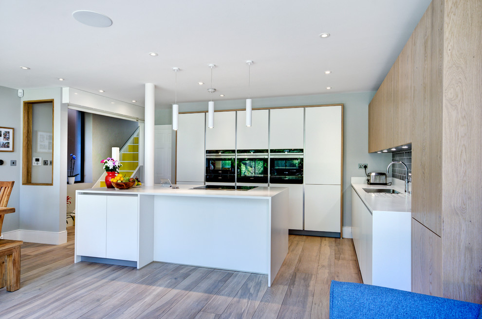 This is an example of a mid-sized contemporary l-shaped open plan kitchen in Berkshire with a double-bowl sink, flat-panel cabinets, white cabinets, solid surface benchtops, grey splashback, subway tile splashback, panelled appliances and with island.