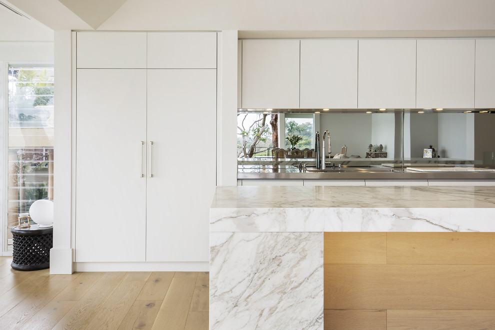 Photo of a large modern single-wall open plan kitchen in Sydney with an integrated sink, flat-panel cabinets, beige cabinets, marble benchtops, metallic splashback, mirror splashback, black appliances, light hardwood floors and with island.