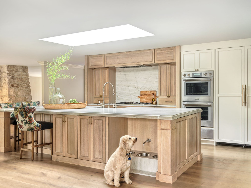Large transitional l-shaped open plan kitchen in St Louis with a farmhouse sink, flat-panel cabinets, white cabinets, quartz benchtops, multi-coloured splashback, panelled appliances, light hardwood floors, with island, brown floor, white benchtop and wallpaper.