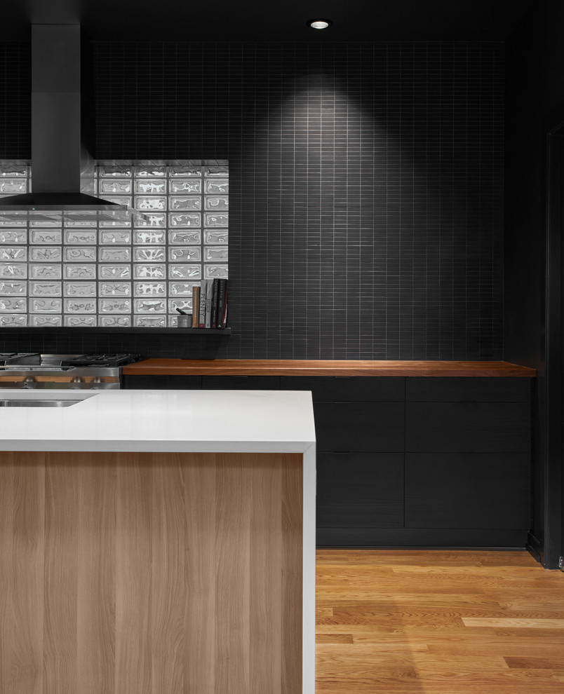 Example of a mid-sized trendy single-wall medium tone wood floor eat-in kitchen design in Chicago with black backsplash, stainless steel appliances, an island, flat-panel cabinets, black cabinets, an undermount sink, wood countertops and glass tile backsplash