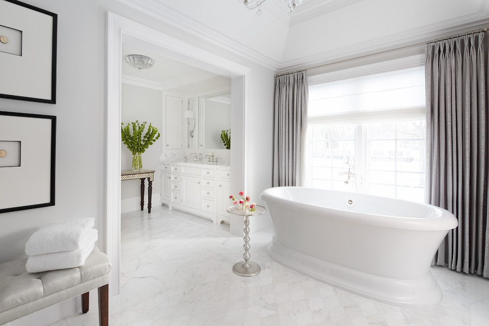 Photo of a large traditional master bathroom in Chicago with white cabinets, a freestanding tub, grey walls, marble floors, recessed-panel cabinets, white tile, stone tile, an undermount sink, marble benchtops and white floor.