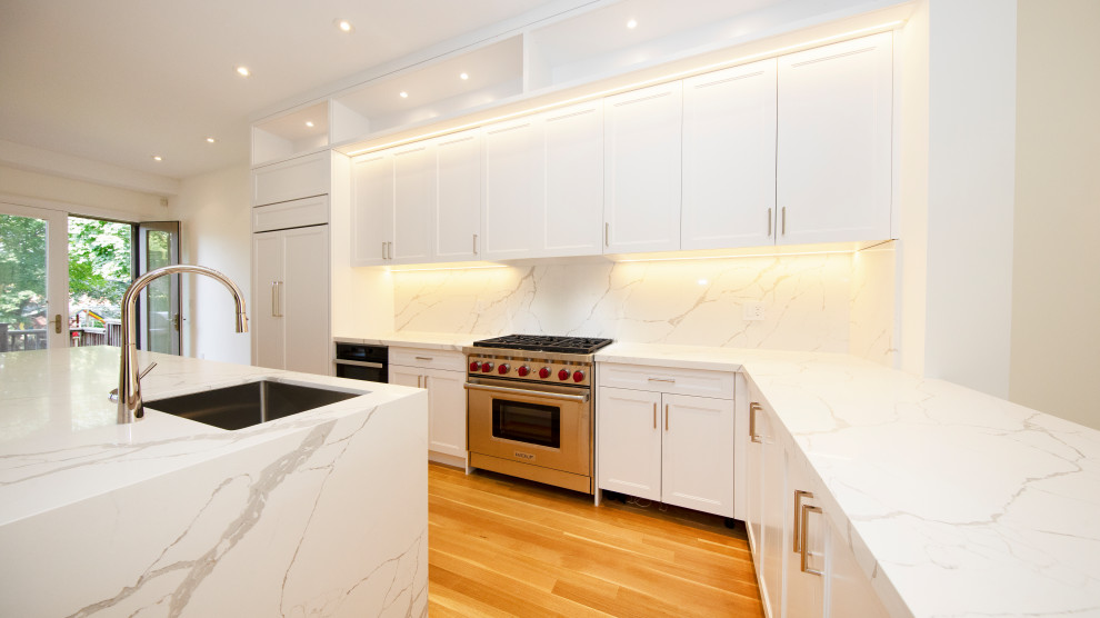 Photo of a large traditional l-shaped kitchen/diner in Toronto with a single-bowl sink, shaker cabinets, white cabinets, engineered stone countertops, white splashback, engineered quartz splashback, integrated appliances, light hardwood flooring, an island and white worktops.