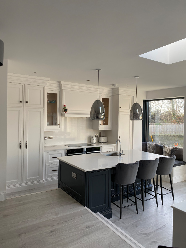 Inspiration for a medium sized farmhouse l-shaped kitchen/diner in Dublin with a built-in sink, shaker cabinets, white cabinets, limestone worktops, beige splashback, ceramic splashback, integrated appliances, laminate floors, an island, grey floors, beige worktops and a vaulted ceiling.