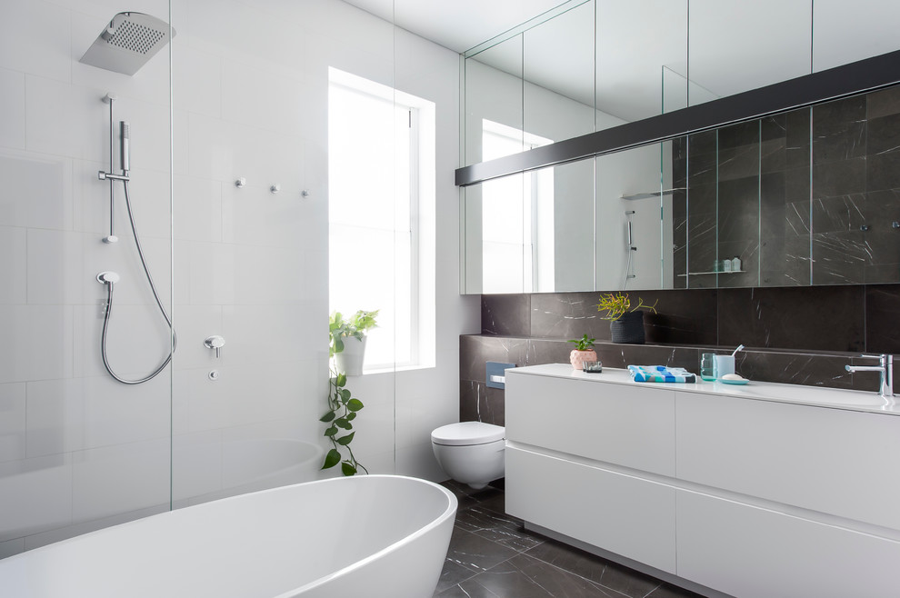Inspiration for a mid-sized contemporary 3/4 bathroom in Sydney with furniture-like cabinets, white cabinets, a freestanding tub, an open shower, a wall-mount toilet, gray tile, stone tile, white walls, marble floors, an integrated sink and solid surface benchtops.