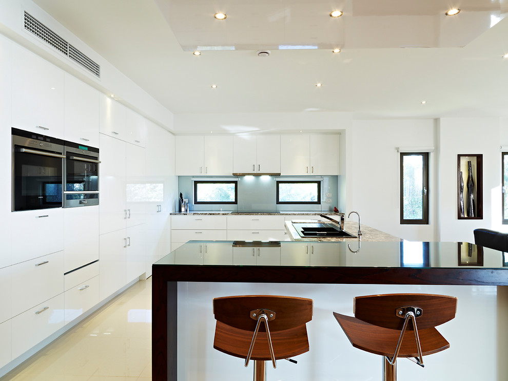 This is an example of a contemporary l-shaped open plan kitchen in Melbourne with a double-bowl sink, flat-panel cabinets, white cabinets, grey splashback, black appliances, porcelain floors, with island and beige floor.