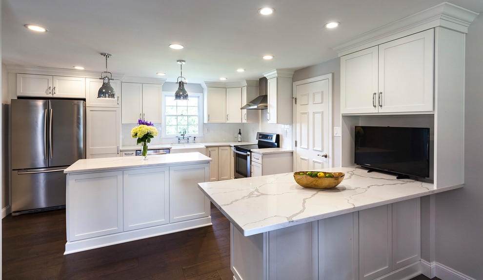 Example of a transitional medium tone wood floor and brown floor kitchen design with a farmhouse sink, white cabinets, quartz countertops, white backsplash, ceramic backsplash, stainless steel appliances and an island