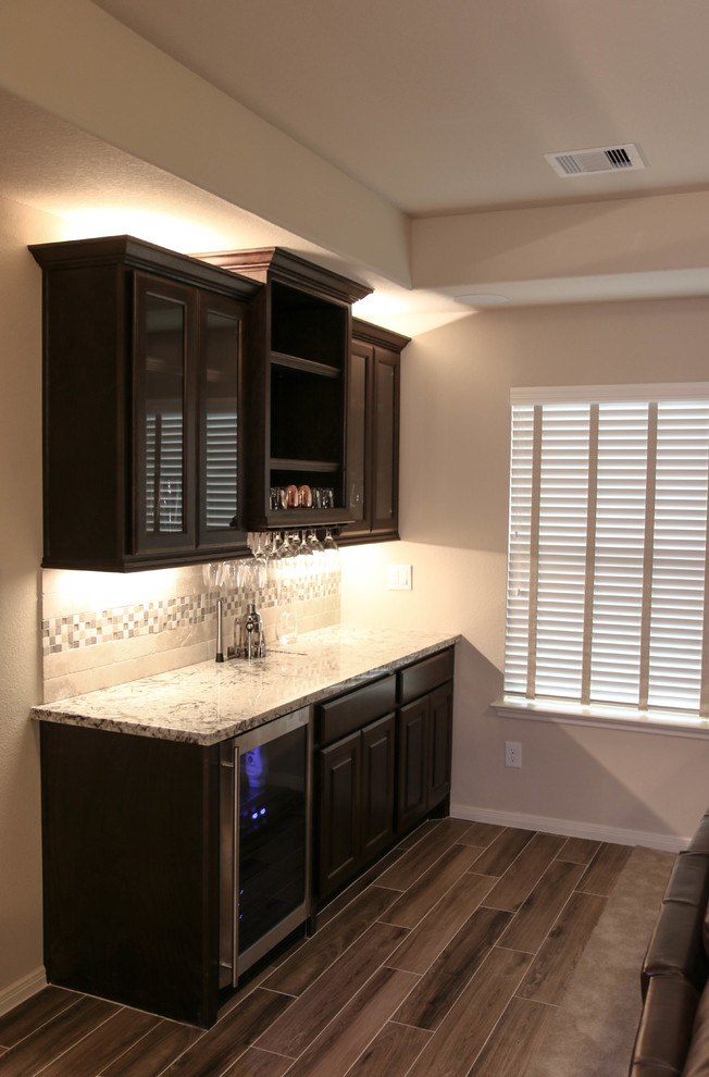 This is an example of a mid-sized transitional single-wall wet bar in Houston with no sink, raised-panel cabinets, dark wood cabinets, granite benchtops, grey splashback, ceramic splashback, porcelain floors and brown floor.
