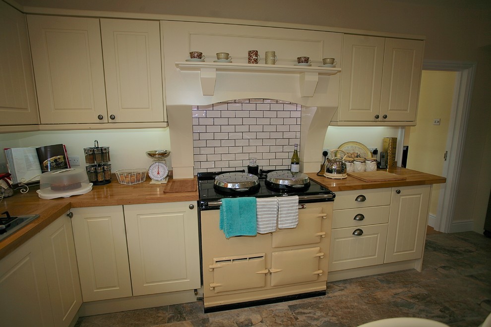 Photo of a large country l-shaped eat-in kitchen in Dorset with a double-bowl sink, beaded inset cabinets, beige cabinets, wood benchtops, white splashback, ceramic splashback, ceramic floors, with island and brown floor.