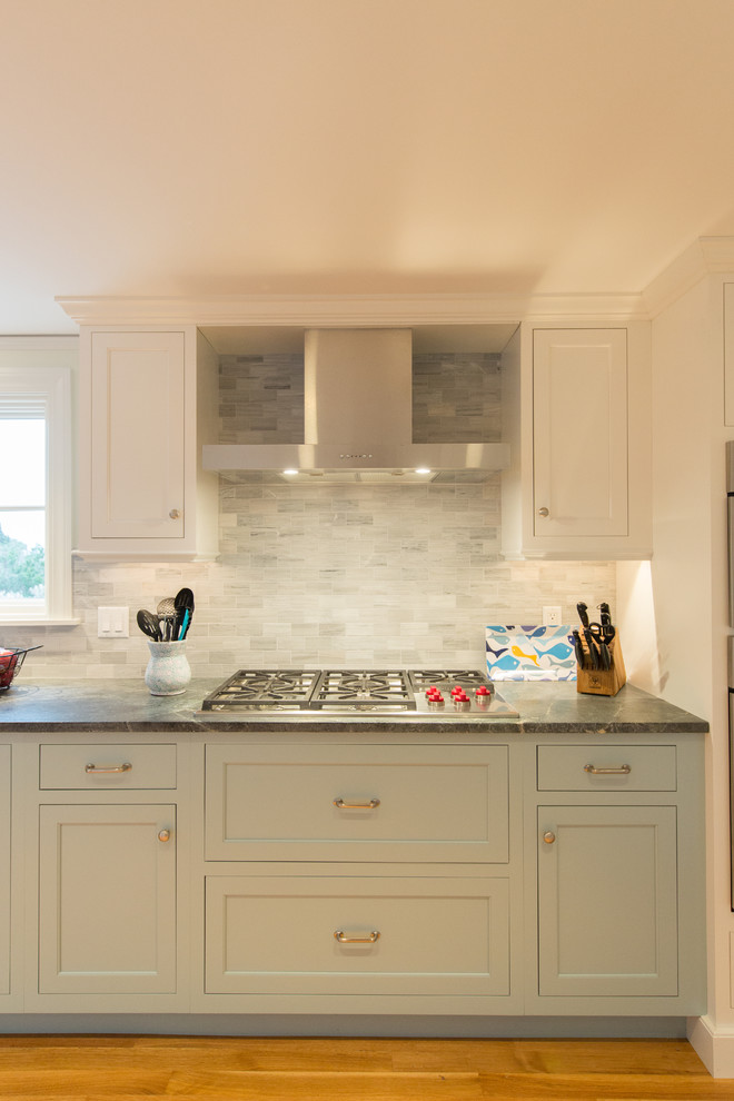 This is an example of a mid-sized beach style kitchen in Boston with a farmhouse sink, shaker cabinets, white cabinets, soapstone benchtops, grey splashback, stone tile splashback, stainless steel appliances, medium hardwood floors, a peninsula and black benchtop.