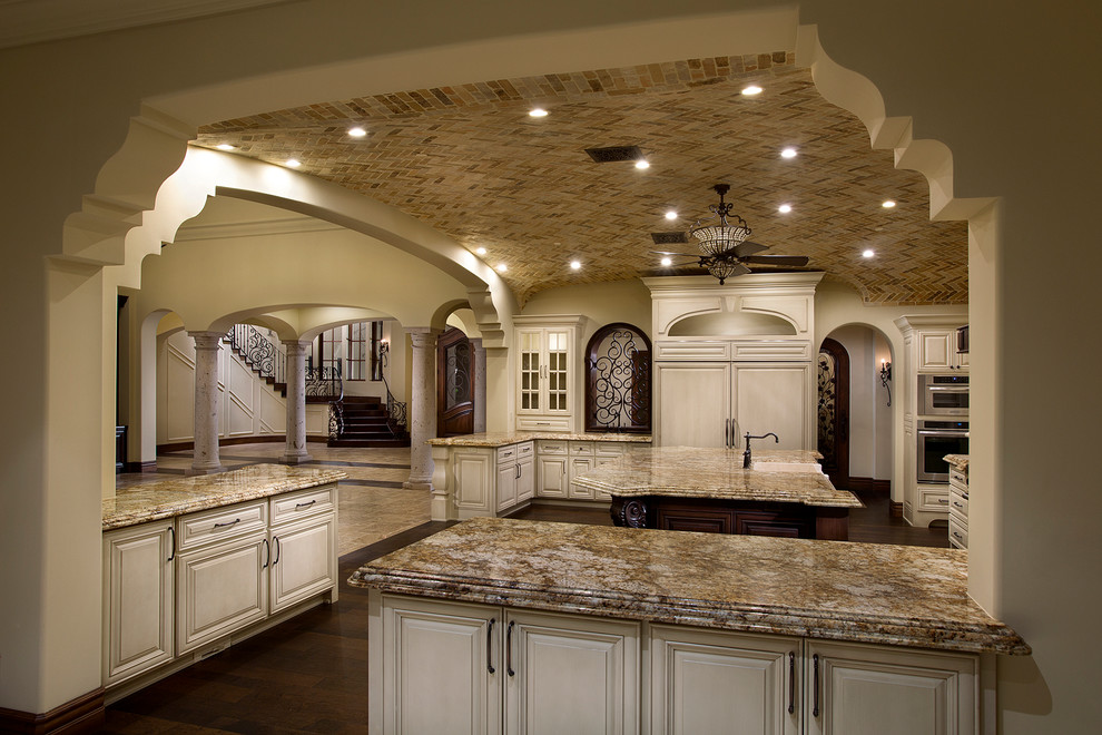This is an example of an expansive mediterranean u-shaped separate kitchen in Phoenix with a farmhouse sink, white cabinets, granite benchtops, beige splashback, mosaic tile splashback, panelled appliances, dark hardwood floors, multiple islands and raised-panel cabinets.