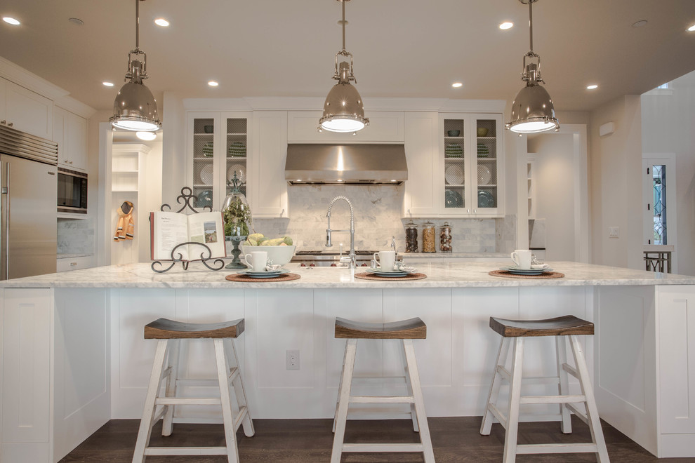 This is an example of an expansive country l-shaped open plan kitchen in Seattle with a farmhouse sink, shaker cabinets, white cabinets, marble benchtops, white splashback, stone tile splashback, stainless steel appliances, dark hardwood floors and with island.