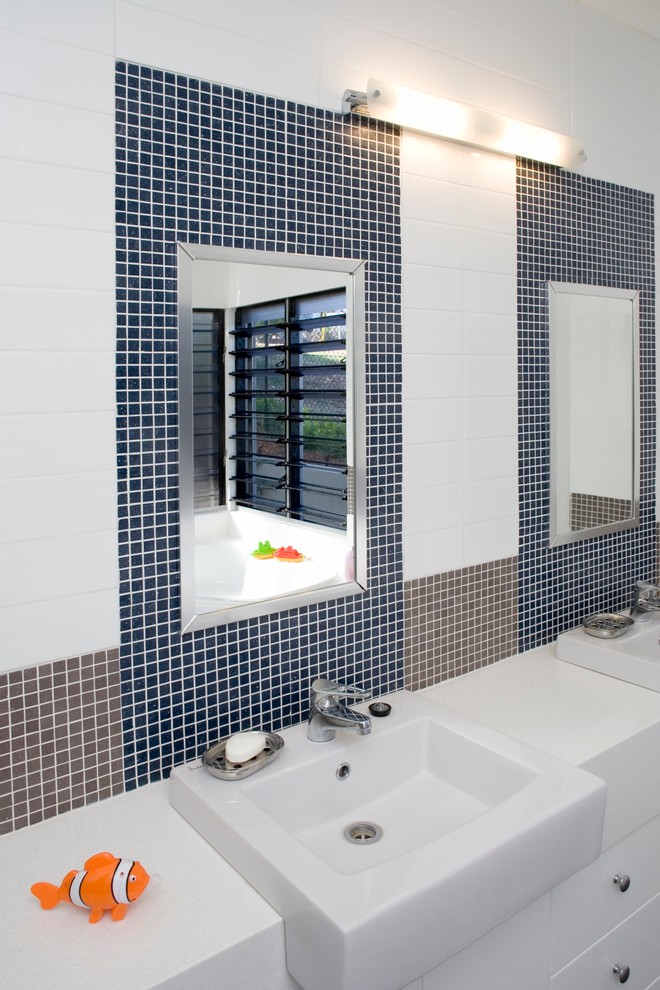 This is an example of a mid-sized tropical kids bathroom in Darwin with a drop-in sink, flat-panel cabinets, white cabinets, solid surface benchtops, a corner tub, multi-coloured tile, mosaic tile and white walls.