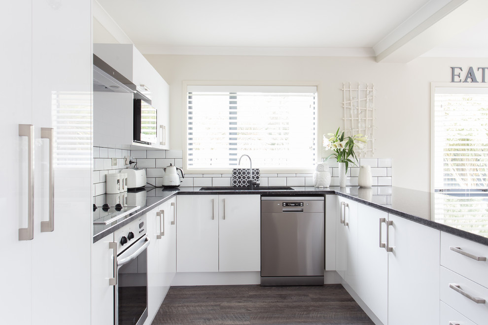 Design ideas for a mid-sized contemporary u-shaped kitchen in Christchurch with an undermount sink, flat-panel cabinets, white cabinets, granite benchtops, white splashback, ceramic splashback, stainless steel appliances, laminate floors, a peninsula, brown floor and black benchtop.