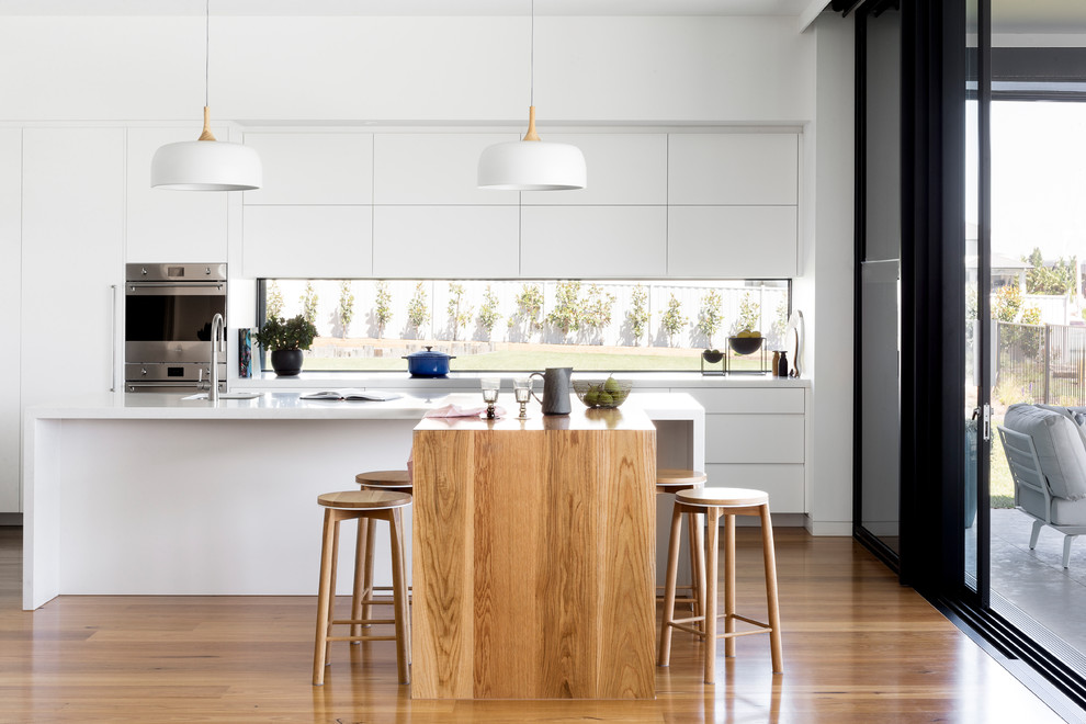 Photo of a contemporary galley kitchen in Melbourne with an undermount sink, flat-panel cabinets, white cabinets, window splashback, stainless steel appliances, medium hardwood floors, brown floor, white benchtop, quartz benchtops and multiple islands.