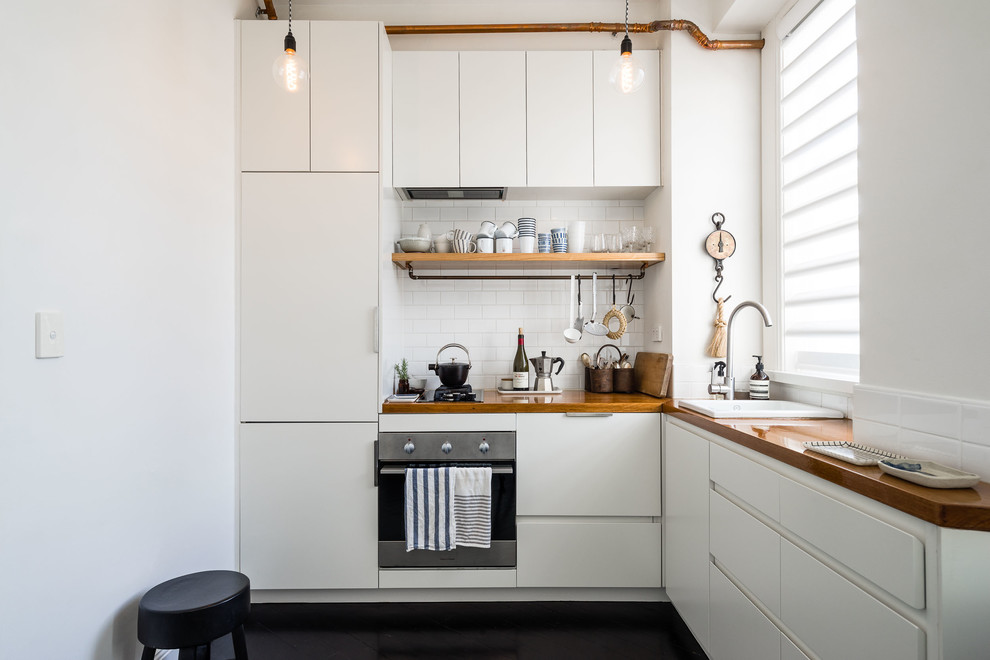 Design ideas for a scandinavian l-shaped kitchen in Sydney with a drop-in sink, flat-panel cabinets, white cabinets, wood benchtops, white splashback, subway tile splashback, black appliances, black floor and brown benchtop.