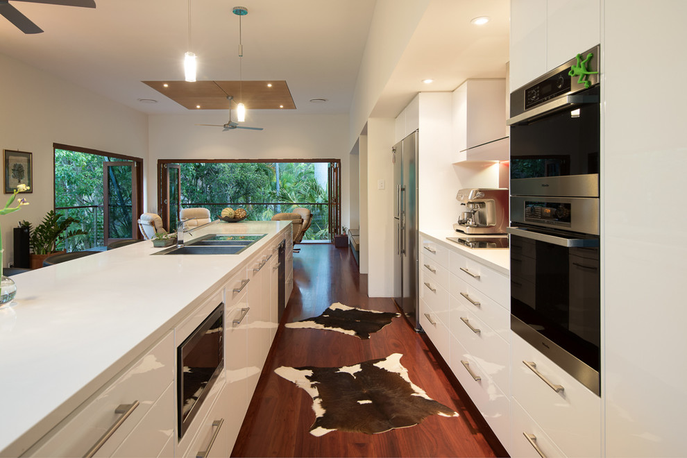 This is an example of a large tropical galley open plan kitchen in Brisbane with a double-bowl sink, flat-panel cabinets, white cabinets, red splashback, stainless steel appliances, dark hardwood floors, with island and brown floor.