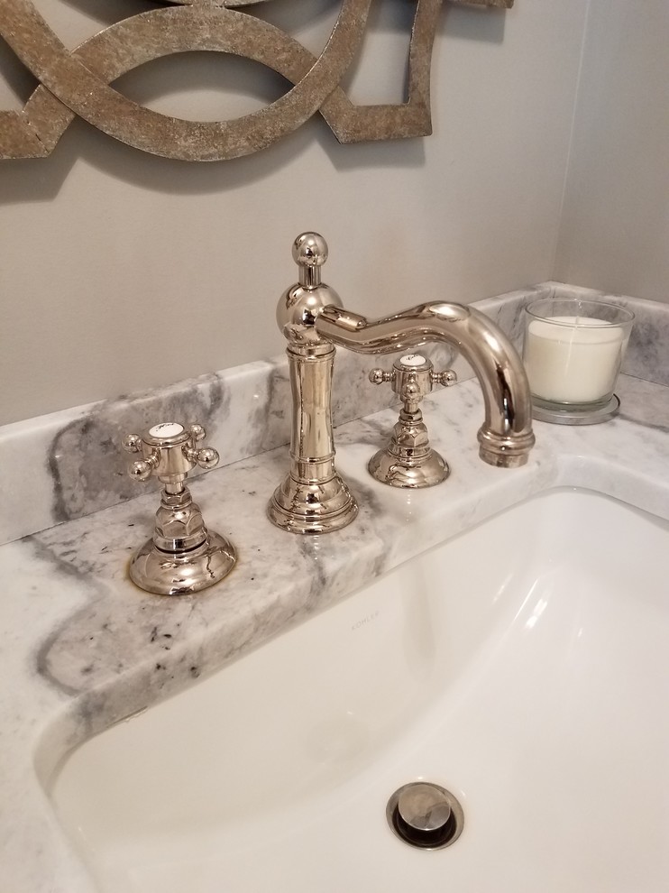 This is an example of a small transitional powder room in New York with recessed-panel cabinets, distressed cabinets, a one-piece toilet, grey walls, laminate floors, an undermount sink, marble benchtops and grey floor.