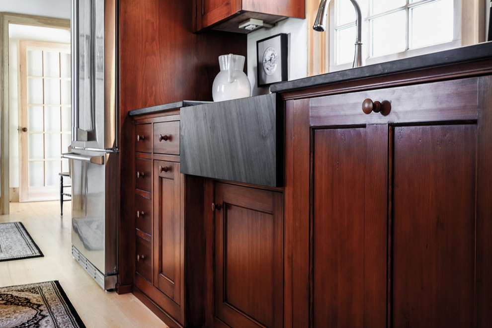 This is an example of a small country l-shaped separate kitchen in Portland Maine with a farmhouse sink, stainless steel appliances, light hardwood floors, a peninsula, recessed-panel cabinets, dark wood cabinets, soapstone benchtops and red splashback.
