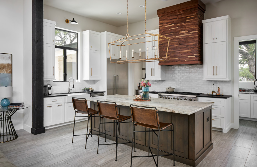 Photo of a transitional u-shaped kitchen in Austin with a farmhouse sink, shaker cabinets, white cabinets, white splashback, subway tile splashback, stainless steel appliances, with island, grey floor and black benchtop.