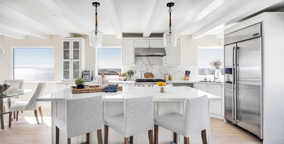 This is an example of a mid-sized beach style l-shaped eat-in kitchen in Los Angeles with shaker cabinets, white cabinets, marble benchtops, white splashback, marble splashback, stainless steel appliances, light hardwood floors, with island, white benchtop, an undermount sink and beige floor.