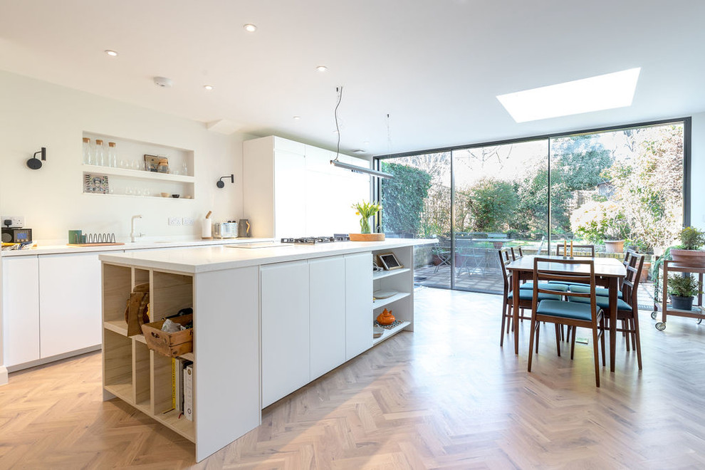 Photo of a mid-sized contemporary single-wall eat-in kitchen in London with flat-panel cabinets, white cabinets, solid surface benchtops, light hardwood floors, with island, white benchtop, an integrated sink, white splashback, panelled appliances and brown floor.