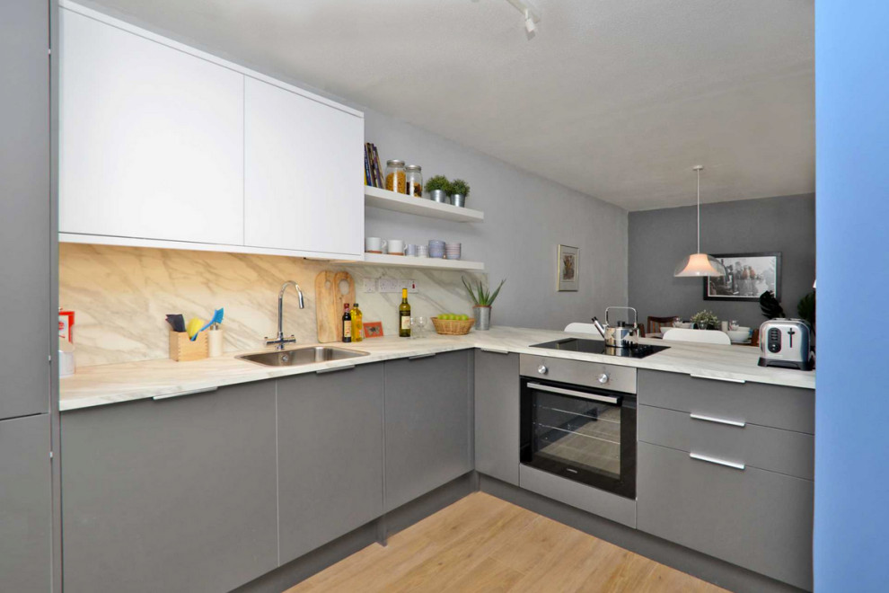 Photo of a medium sized scandinavian l-shaped kitchen/diner in London with a built-in sink, flat-panel cabinets, grey cabinets, laminate countertops, white splashback, integrated appliances, light hardwood flooring, brown floors and white worktops.