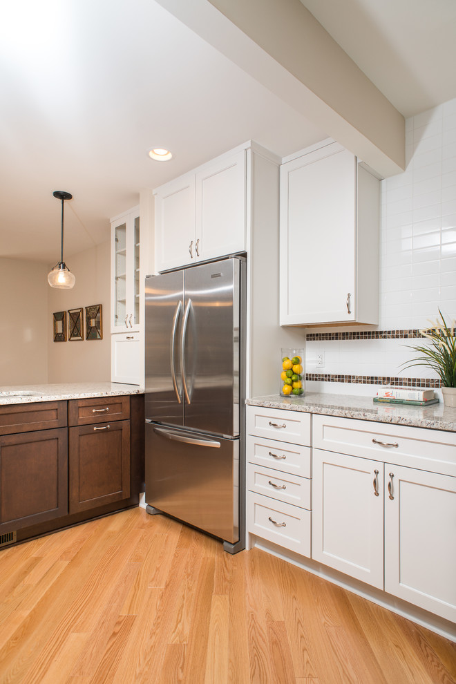 Photo of a mid-sized arts and crafts l-shaped kitchen pantry in Detroit with stainless steel appliances, white cabinets, granite benchtops, white splashback, porcelain splashback, light hardwood floors, a peninsula and shaker cabinets.