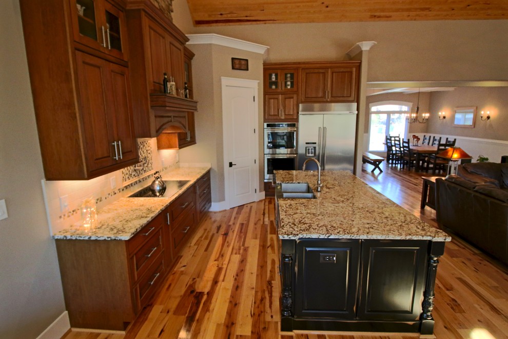 Example of a mid-sized transitional l-shaped light wood floor and beige floor kitchen pantry design in Raleigh with a farmhouse sink, raised-panel cabinets, medium tone wood cabinets, granite countertops, multicolored backsplash, glass tile backsplash, stainless steel appliances, an island and brown countertops