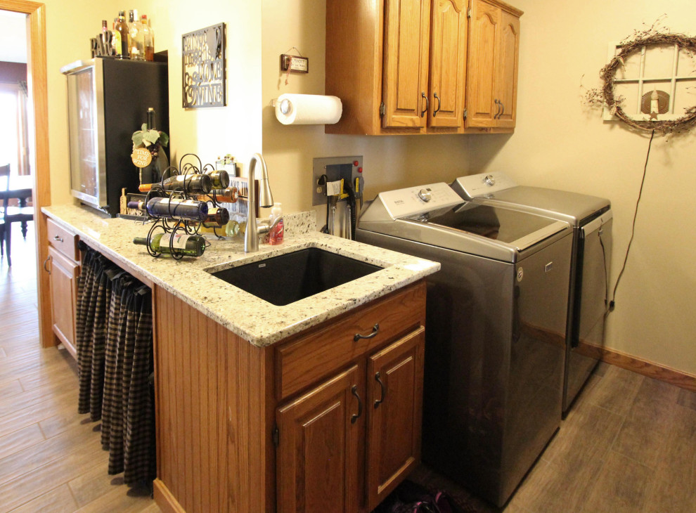 This is an example of a large country l-shaped laundry room in Columbus with an undermount sink, raised-panel cabinets, medium wood cabinets, granite benchtops, yellow walls, porcelain floors, brown floor and beige benchtop.