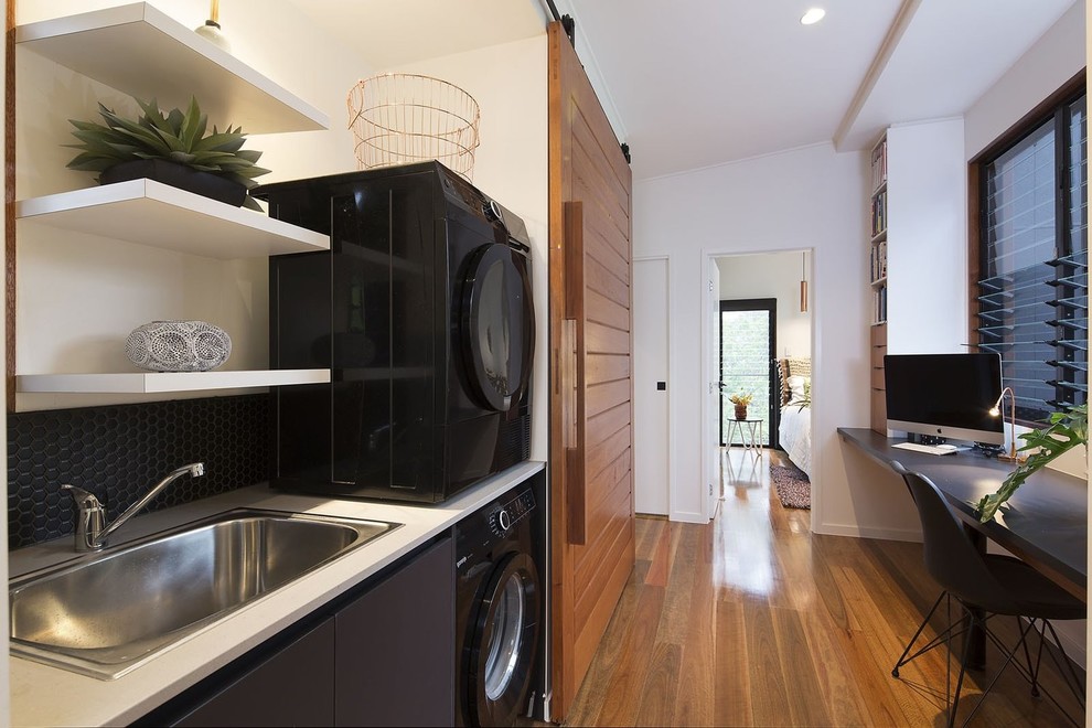 Photo of a modern single-wall dedicated laundry room in Brisbane with flat-panel cabinets, black cabinets, quartz benchtops, white walls, medium hardwood floors, a stacked washer and dryer, brown floor and white benchtop.
