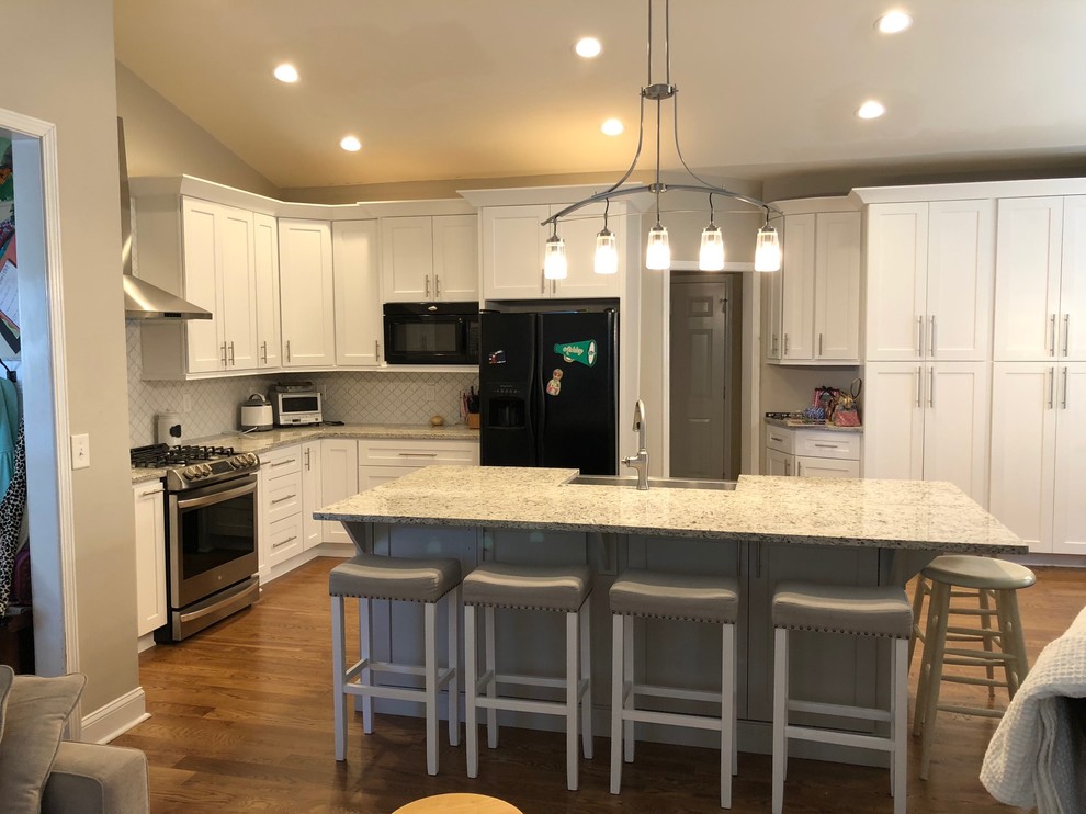 Photo of a mid-sized modern l-shaped eat-in kitchen in Atlanta with a farmhouse sink, shaker cabinets, white cabinets, granite benchtops, white splashback, ceramic splashback, stainless steel appliances, medium hardwood floors, with island and brown floor.