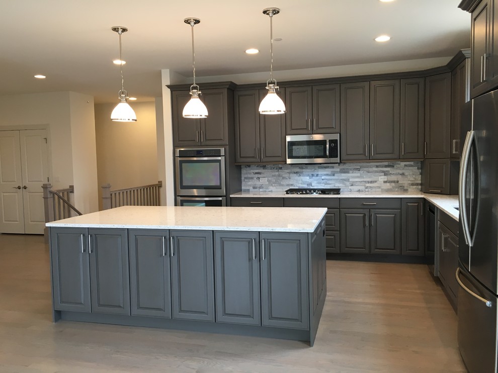 Transitional l-shaped open plan kitchen in Chicago with a double-bowl sink, raised-panel cabinets, grey cabinets, quartz benchtops, grey splashback, stone tile splashback, stainless steel appliances, light hardwood floors, with island and grey floor.