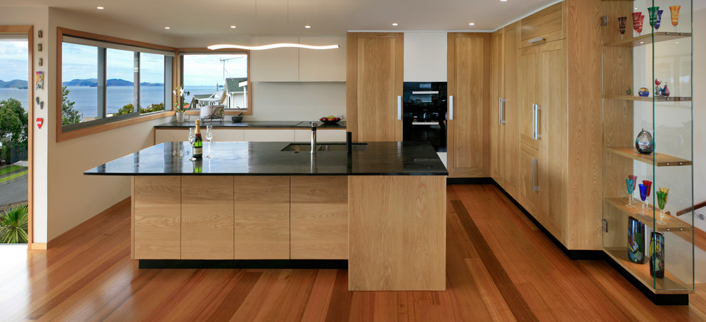 Photo of a large modern l-shaped open plan kitchen in Auckland with a double-bowl sink, recessed-panel cabinets, medium wood cabinets, granite benchtops, black appliances, medium hardwood floors, with island and black benchtop.
