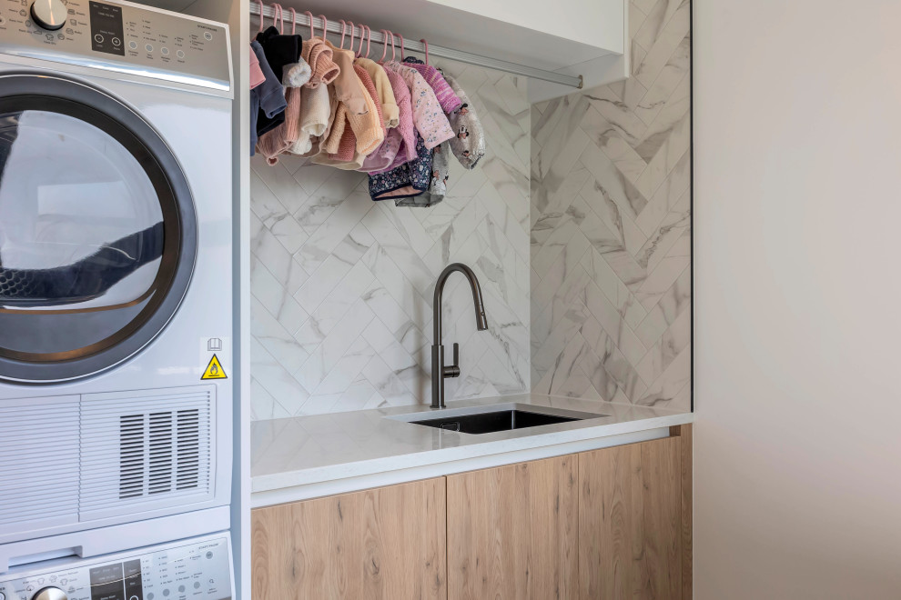 Inspiration for a small modern single-wall dedicated laundry room in Auckland with an undermount sink, flat-panel cabinets, medium wood cabinets, quartz benchtops, white splashback, ceramic splashback, white walls, porcelain floors, a stacked washer and dryer, grey floor and white benchtop.