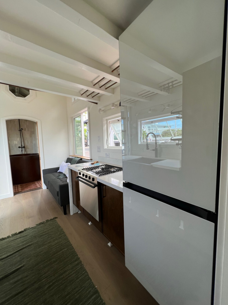 Medium sized galley kitchen in Hawaii with glass-front cabinets, brown cabinets, engineered stone countertops, white splashback, white appliances, light hardwood flooring, no island, beige floors, white worktops and exposed beams.