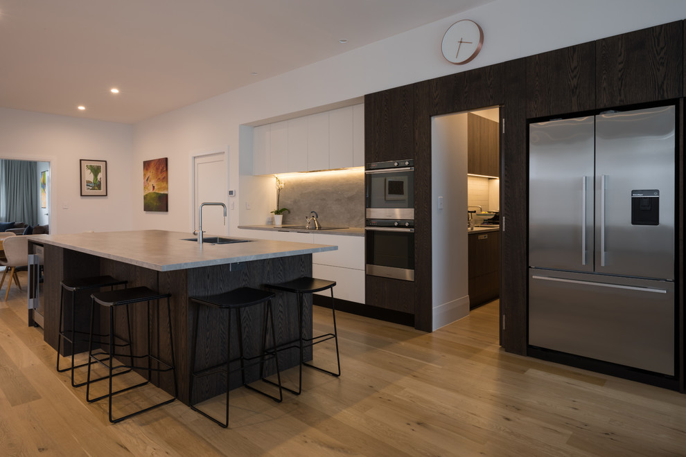 This is an example of a large contemporary single-wall eat-in kitchen in Auckland with a double-bowl sink, flat-panel cabinets, dark wood cabinets, solid surface benchtops, grey splashback, stone slab splashback, stainless steel appliances, light hardwood floors, with island, brown floor and grey benchtop.