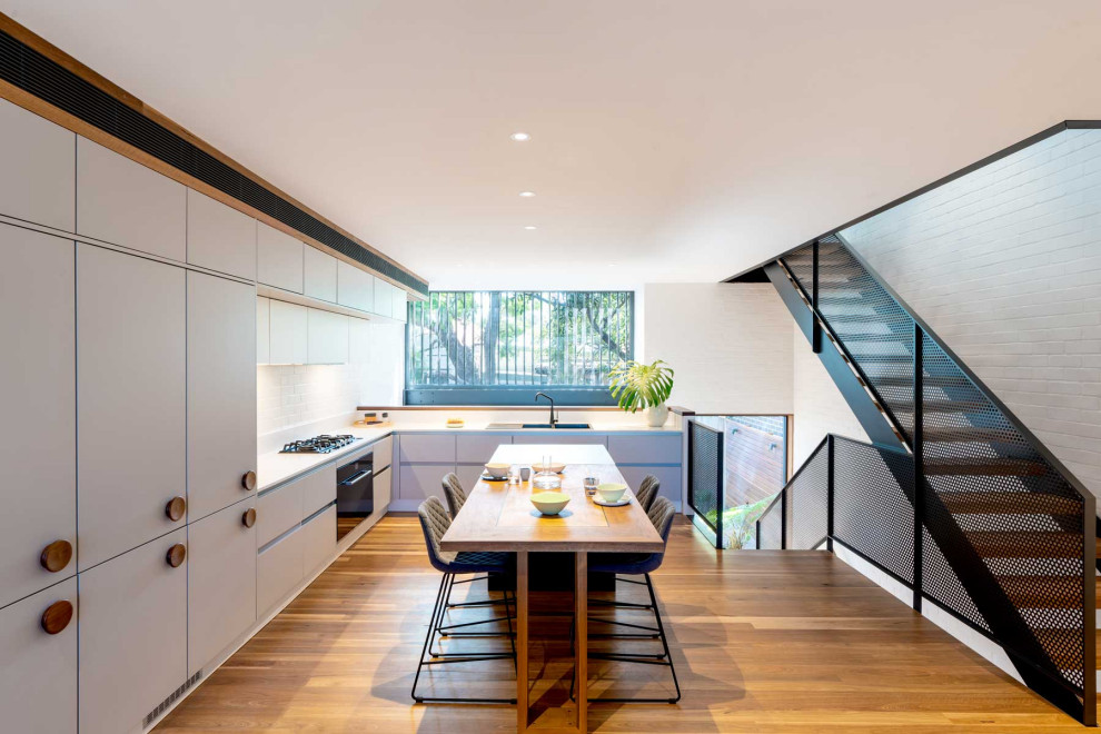 Large contemporary l-shaped eat-in kitchen in Sydney with a double-bowl sink, flat-panel cabinets, white cabinets, white splashback, panelled appliances, no island, brown floor and white benchtop.