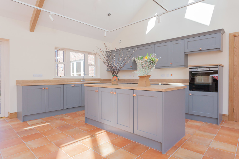 This is an example of a mid-sized contemporary l-shaped open plan kitchen in Belfast with a drop-in sink, recessed-panel cabinets, grey cabinets, wood benchtops, brown splashback, timber splashback, stainless steel appliances, ceramic floors, with island, orange floor and brown benchtop.