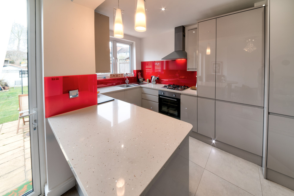 Mid-sized modern galley eat-in kitchen in London with an integrated sink, flat-panel cabinets, red cabinets, granite benchtops, red splashback, glass sheet splashback, panelled appliances, travertine floors, with island, beige floor and multi-coloured benchtop.