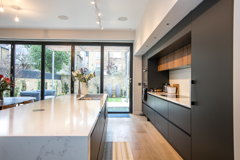 Photo of a mid-sized contemporary galley open plan kitchen in London with a double-bowl sink, flat-panel cabinets, grey cabinets, quartz benchtops, white splashback, stainless steel appliances, medium hardwood floors, with island and white benchtop.