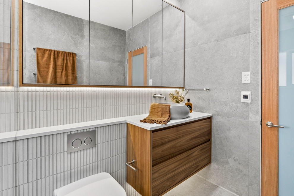 Exemple d'une salle de bain principale tendance en bois brun de taille moyenne avec un placard en trompe-l'oeil, une douche ouverte, WC à poser, un carrelage gris, des carreaux de porcelaine, un mur gris, un sol en carrelage de porcelaine, une vasque, un plan de toilette en surface solide, un sol gris, aucune cabine, un plan de toilette blanc, meuble simple vasque et meuble-lavabo encastré.