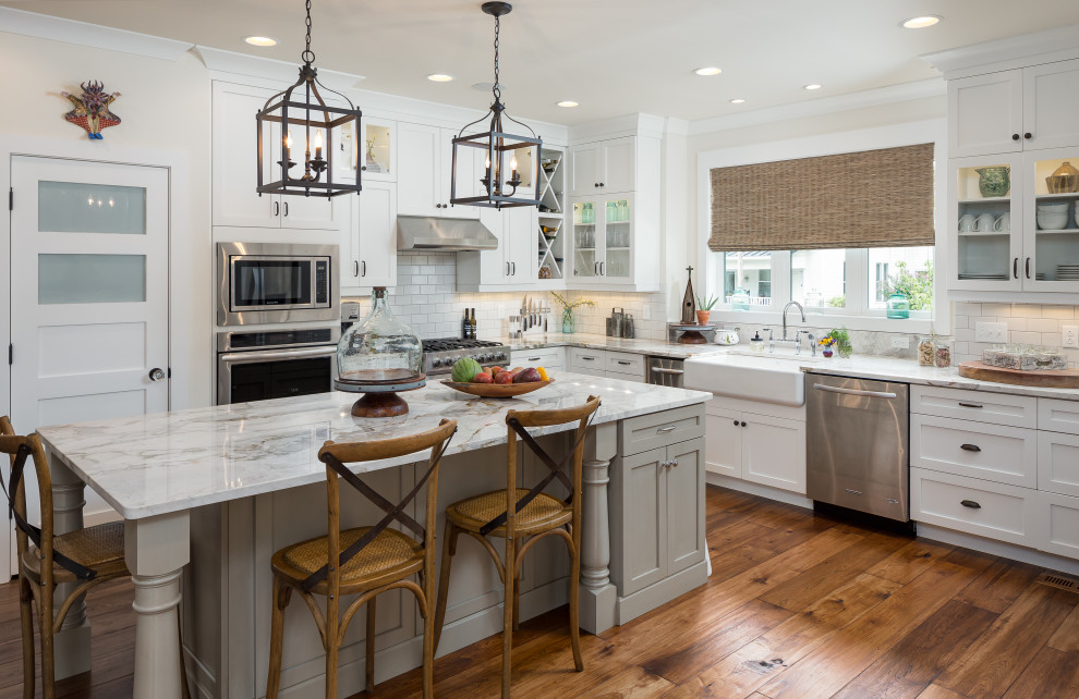 Inspiration for a country l-shaped kitchen in Nashville with a farmhouse sink, shaker cabinets, white cabinets, white splashback, subway tile splashback, stainless steel appliances, medium hardwood floors, with island, brown floor and white benchtop.